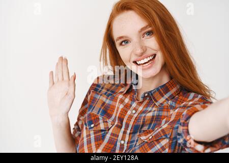 Nahaufnahme einer jungen Frau mit roten langen Haaren und blauen Augen, die Selfie gemacht und Hi. gesagt hat Mädchen blogger winkt Hand und lächelt an der Kamera Stockfoto