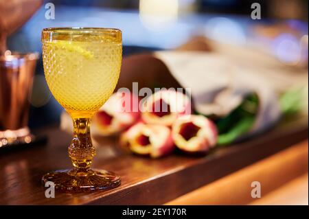 Gelber Cocktail mit Zitrone und Eis in einem schönen mittelalterlichen Glas neben einem Bouquet von gelb-roten Tulpen. Internationaler Frauentag Stockfoto