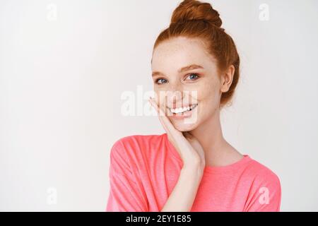 Schönheit und Hautpflege. Lächelndes Mädchen mit Ingwer-Haar gekämmt in chaotischen Brötchen, berühren perfekte Haut und lächeln, stehen ohne Make-up auf weißem Hintergrund Stockfoto