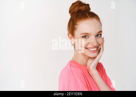 Schönheit und Hautpflege. Lächelndes Mädchen mit Ingwer-Haar gekämmt in chaotischen Brötchen, berühren perfekte und leuchtende Haut und lächelnd, zeigt Gesicht ohne Make-up Stockfoto