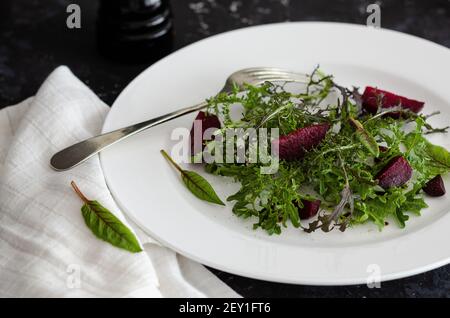 Grüner und lila mizuna und Rote Beete Salat auf weißem Teller, mit Gabel und weißer Serviette, auf schwarzem Hintergrund. Stockfoto