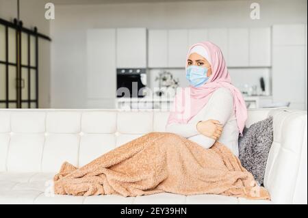 Kranke muslimische Frau trägt medizinische Maske bedeckt Decke allein auf der Couch zu Hause sitzen, fühlt Grippe und Fieber. Selbstisolationskonzept, Coronavirus-Pandemie Stockfoto