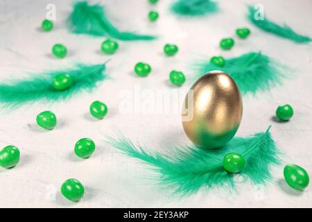 Goldenes Ei auf weißem Betongrund mit grünen Federn und grünen Pralinen in Form eines Eies. Frohe Ostern Konzept. Weichfokus Stockfoto