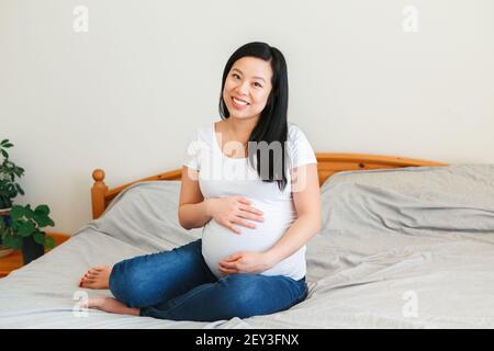 Lächelnd asiatische chinesische schwangere Frau sitzt auf dem Bett berühren Bauch. Junge Dame in weißen T-Shorts und blauen Jeans zu Hause erwartet. Gesunde glückliche Eltern Stockfoto