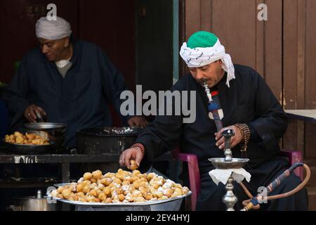 Porträt eines ägyptischen Mannes, der eine Shisha-Pfeife raucht und frittierte Süßigkeiten in einem Café (ahwa) im islamischen Viertel von Kairo, Ägypten, isst Stockfoto