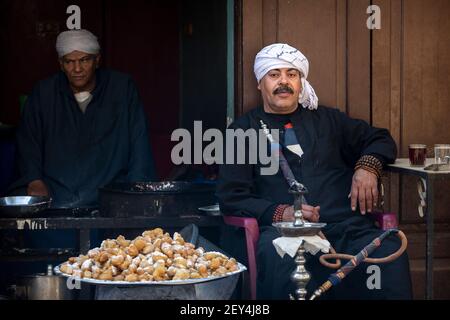 Porträt eines ägyptischen Mannes, der in einem Café (ahwa) im Islamischen Viertel von Kairo, Ägypten, neben einer Schale mit frittierten Süßigkeiten eine Shisha-Pfeife raucht Stockfoto