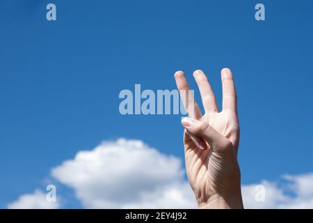 Nahaufnahme Geste einer Frau Hand zeigt drei Finger isoliert auf einem Hintergrund von blauen Himmel mit Wolken, Symbol Zeichen Sprache Nummer drei. Stockfoto