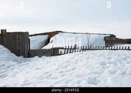 Altes Dorfhaus, alter Holzzaun, der während eines Schneesturms mit Schnee bedeckt ist. Das Haus ist nach einem Sturm oder starkem Schneefall mit Schnee bedeckt Stockfoto