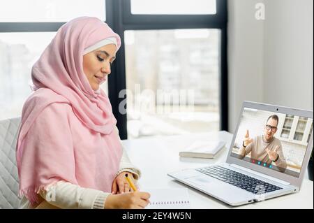 Islamische Studentin, die online studiert, nimmt Notizen, während männliche Tutorin, Lehrerin, die auf dem Laptop-Bildschirm spricht. Muslimische Frau trägt Hijab beobachten Webinar, Online-Training und Schreiben mit Stift, E-Studie Stockfoto