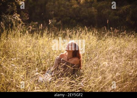 Friedliche junge Frau, die während auf dem Feld im Gras sitzt Sommer Stockfoto