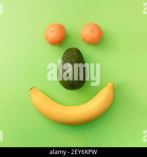 Positives Smiley-Gesicht aus Banane, Avocado und Mandarine auf grünem Hintergrund. Flaches Lay buntes Konzept. Stockfoto