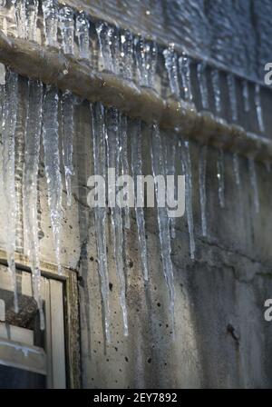 Gefrorene spitzen Eiszapfen, die an einem industriellen Metallrohr auf grunbendem Wasser hängen, beschädigten im Hintergrund Eisschatten auf der Betonaußenwand Stockfoto