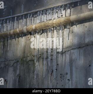 Gefrorene spitzen Eiszapfen, die an einem industriellen Metallrohr auf grunbendem Wasser hängen, beschädigten im Hintergrund Eisschatten auf der Betonaußenwand Stockfoto