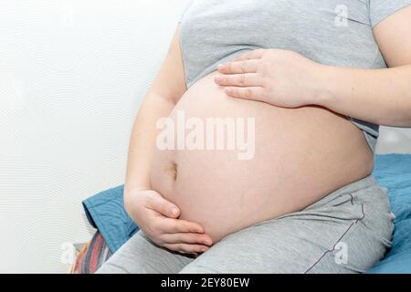 Ein schwangeres Mädchen sitzt zu Hause auf dem Bett und hält ihre Hände auf dem Bauch. Schwangerschaft und Familienkonzept. Stockfoto