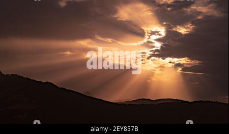 Lichtstrahlen, die durch die Wolken in einem wunderschönen Sonnenuntergang oder Sonnenaufgang hinter der Silhouette des Berges filtern. Malerischer Blick auf den Himmel mit Sonnenstrahlen, wunderschön Stockfoto