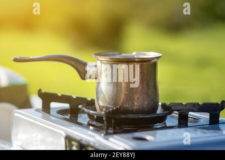 Tee und Kaffee machen mit einem tragbaren Gasherd auf naturgrünem Hintergrund im Ferienlager an sonnigen Tagen. Campingkocher mit kleiner Kaffeekanne im Freien. Stockfoto