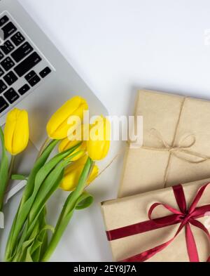 Grauer Laptop, Bouquet von gelben Tulpen und eingewickelte Geschenke auf weißem Tisch. Flatlay Stockfoto