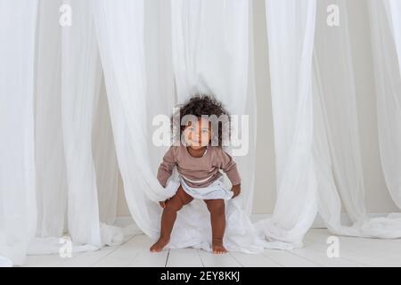 Kleine lockige afroamerikanische Mädchen hat Spaß lacht Tänze zwischen luftigen weißen Tüll Stoff. Minimalistische Einrichtung, skandinavischer Stil. Frohe Kindheit. Der Stockfoto