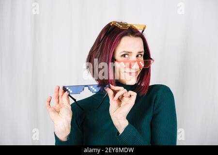 Lustige Frau Versucht Viele Farbe Pixel Brillen. Young Happy Girl mit lila Haaren tragen viele bunte Brille auf grauem Hintergrund Stockfoto