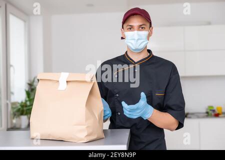 Junger Mann in medizinischen Handschuhen und Schutzmaske halten Papiertüten in den Händen. Mann Kurier mit Einkaufstasche. Sichere Online-Lieferung von Stockfoto