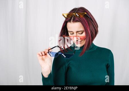 Lustige Frau Versucht Viele Farbe Pixel Brillen. Young Happy Girl mit lila Haaren tragen viele bunte Brille auf grauem Hintergrund Stockfoto