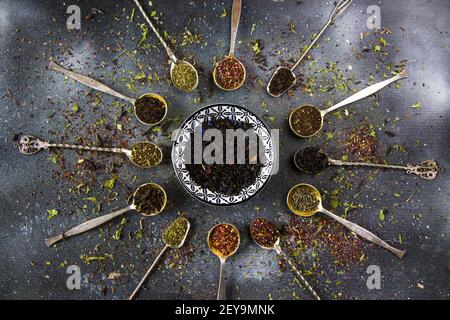 Flache Lay einer Auswahl von getrockneten Teeblättern in Eine keramische Untertasse und vintage Besteck Löffel auf einem dunklen Hintergrund Stockfoto