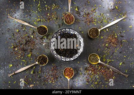 Flache Lay einer Auswahl von getrockneten Teeblättern in Eine keramische Untertasse und vintage Besteck Löffel auf einem dunklen Hintergrund Stockfoto