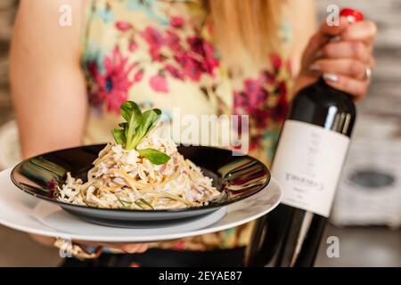 Nahaufnahme des mittleren Abschnitts einer unbekannten kaukasischen Frau mit einem Teller gekochte Carbonara Pasta Spaghetti in einem Teller mit Flasche Wein neben zu Hause - Stockfoto