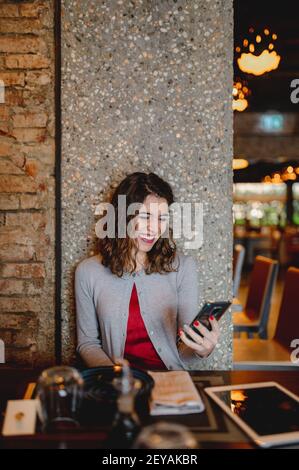 Eine schöne lachende junge Frau, die Spaß mit ihrem Handy hat Im Restaurant an der Wand sitzend Stockfoto