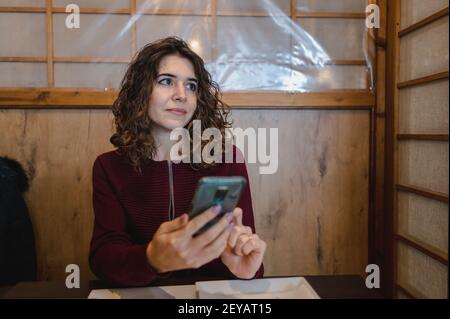 Eine junge lockige Brünette, die in einem Café sitzt und benutzt Ihr Telefon Stockfoto