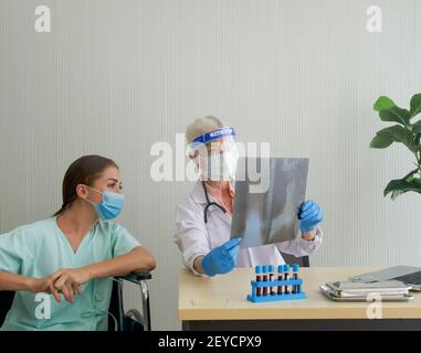 Eine alte Ärztin mit Gesichtsschutz und Schutzmaske zeigt dem im Rollstuhl sitzenden Patienten den Röntgenfilm der Brust. Beschreiben Sie die Symptome o Stockfoto