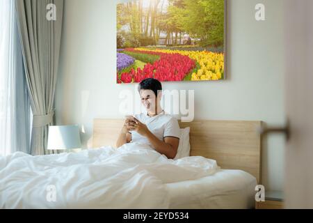 Ein asiatischer Mann sitzt mit einer weißen Decke im Bett und checkt seine E-Mails am Morgen zu Hause auf seinem Handy, bevor er duscht. Stockfoto