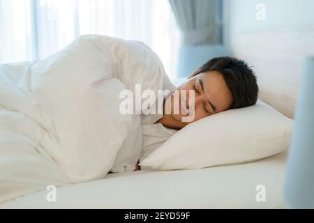 Asiatische Mann schlafen und mit guten Träumen in weißen Decke in den Morgen. Ruhe nach der Arbeit ermüdend im Schlafzimmer zu Hause Stockfoto