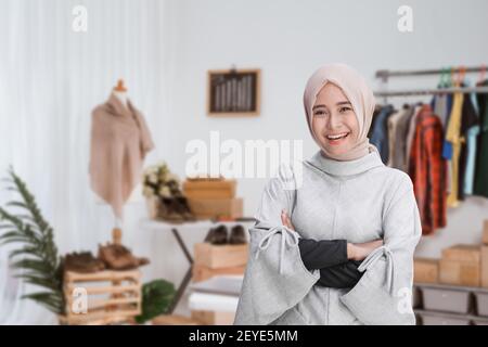 Porträt einer jungen attraktiven muslimischen Frau, die mit gekreuzten Armen steht In einem Zimmer mit Schreibtisch, Schaufensterpuppe und Kleidung im Designer-Arbeitsbereich Stockfoto