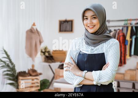 Porträt einer jungen attraktiven muslimischen Frau, die mit gekreuzten Armen steht In einem Zimmer mit Schreibtisch, Schaufensterpuppe und Kleidung im Designer-Arbeitsbereich Stockfoto