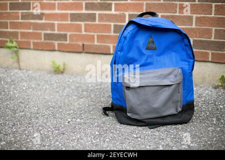 Schulrucksack auf der Straße. Blauer Jungen Rucksack auf dem Bürgersteig in der Nähe der roten Backsteinmauer der Schule im Hintergrund. Zurück zur Schule pädagogisches Konzept. Stockfoto