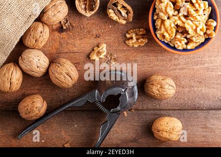 Flachbild zeigt eine gusseiserne Walnusszange (ein Nussknacker-Werkzeug) mit rohen frischen Walnüssen in Schale auf Holztisch. Eine kleine Schüssel mit Walnusskernen Stockfoto