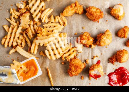 Flach aufgelegtes Bild eines chaotischen Essens mit Waffelfrites und Chicken Nuggets auf Backpapier zusammen mit Ketchup und anderen Saucen und Dips. Einige werden gegessen o Stockfoto
