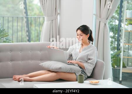 Eine asiatische Frau verbringt Zeit zu Hause, sitzt auf dem Sofa, mit ihrem Laptop Stockfoto