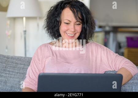 Lächelnde Frau sitzt an ihrem Computer auf der Couch Stockfoto