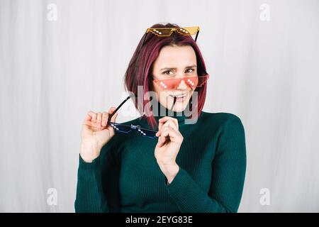 Lustige Frau Versucht Viele Farbe Pixel Brillen. Young Happy Girl mit lila Haaren tragen viele bunte Brille auf grauem Hintergrund Stockfoto