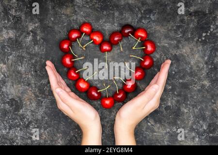 Crop-Ansicht der weiblichen Hände, die vorgeben, ein symbolisches Herz aus roten Kirschen auf einem grungigen Tisch zu halten. Kreatives Pflegekonzept. Stockfoto