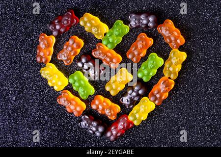 Gummibären in Herzform auf einem schwarzen, mit Zucker überzogenen Hintergrund angeordnet. Stockfoto