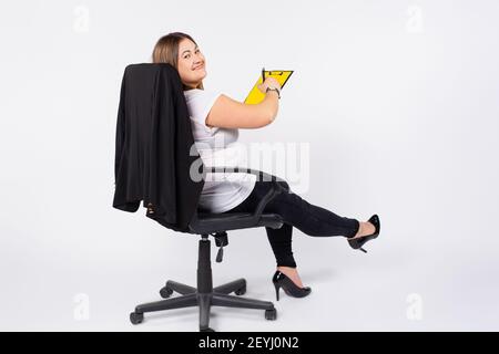 Übergewichtige Frau, die im Büro arbeitet. Ledersessel und gelbe Notizblock. Blickt in die Kamera. Weißer Hintergrund. Seitenraum. Stockfoto