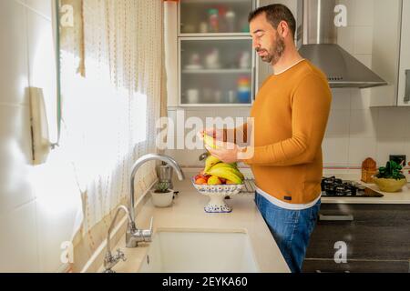 Bärtiger Mann in einem orangefarbenen Pullover, der eine Banane aus der Obstschale in der Küche aufnimmt und analysiert. Gesunde Ernährung Konzept Stockfoto