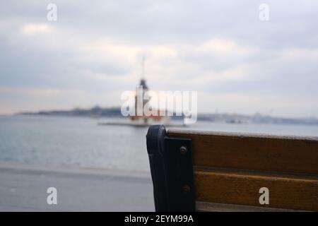 Ecke der Straßenbank aus Holz und Metall zusammen mit verschwommenem Jungfernturm istanbul Hintergrund und bosporus Meer während bewölkten Himmel Hintergrund. Stockfoto