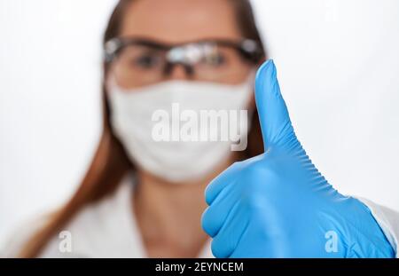 Hand in blauen Schutzhandschuh zeigt Daumen nach oben, verschwommen Frau Gesicht mit Baumwolle Nase Mundmaske und Schutzbrille im Hintergrund Stockfoto
