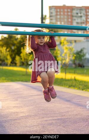 Kleines Mädchen in einem Kleid mit Tupfen zieht sich bei Sonnenuntergang an einer horizontalen Stange im Park hoch. Gesunde Sommeraktivitäten für Kinder. Stockfoto