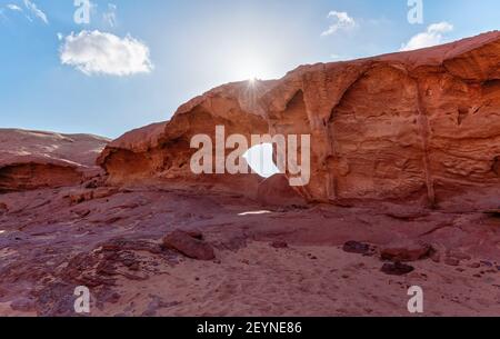 Kleiner Bogen oder kleine Felsenfensterformation in der Wadi Rum Wüste, helle Sonne scheint auf rotem Staub und Felsen, blauer Himmel darüber Stockfoto