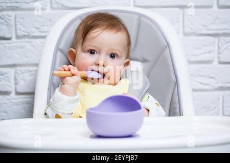 Ein kleines Kind sitzt auf einem Hochstuhl und isst mit einem Löffel Essen von einem Teller. Baby Silikon Utensilien zum Füttern von Babys Stockfoto